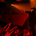 One Ok Rock member stand on the edge of the Algonquin Commons Theatre stage with his hand outreached to the audience while the floor level audience all have their hands up as well. The photo has a red cast over it.