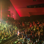 Lead singer from the band Switchfoot walks along the path at the bottom of the main level with his microphone as the audience at the floor and main level are all standing and chearing