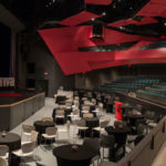 Round tables with linen on them and white chairs are set up on the floor level of the Algonquin Commons Theatre